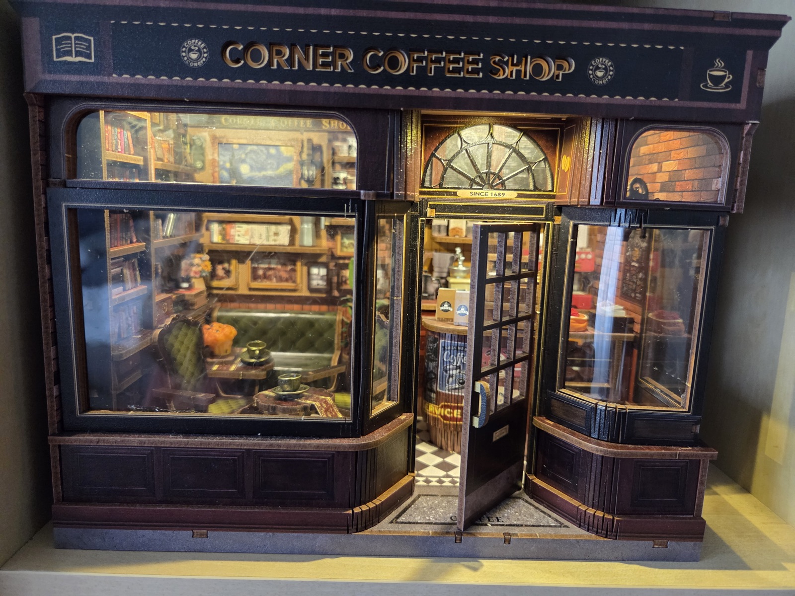 Miniature Corner Coffee Shop room box, lit from inside, with a bookshelf-lined café seen through the shop window and an open door